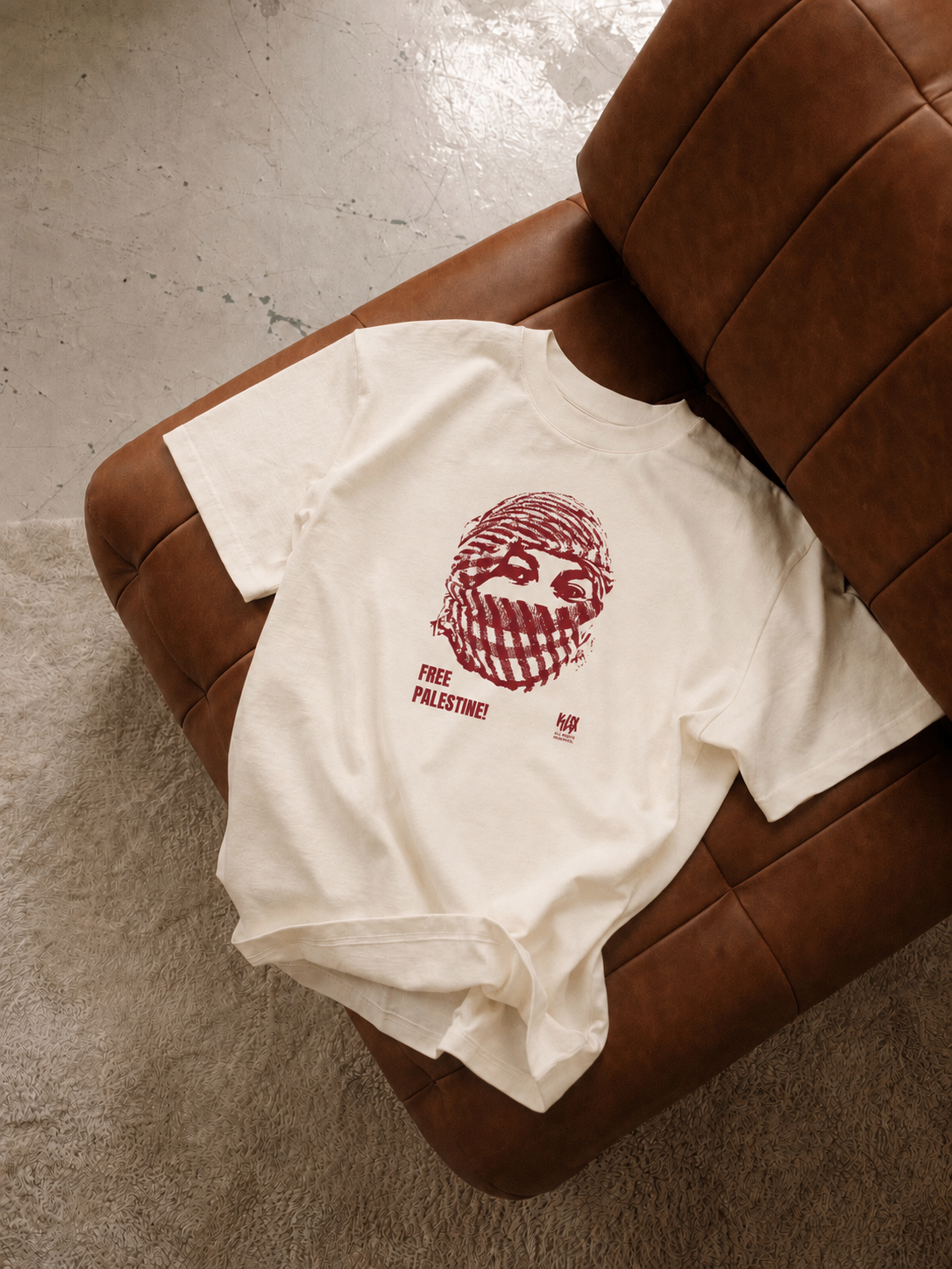 An off-white short-sleeve t-shirt with a dark red graphic print of a face in a striped mask and the text 'FREE PALESTINE' underneath, styled on a brown leather armchair over a concrete floor.